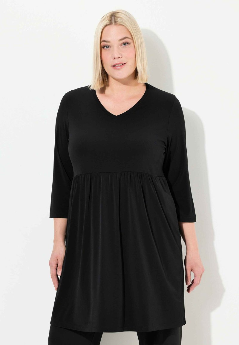 Mustat, polvipituiset tinukat, joissa on V-pääntie ja kolme neljänneshihaa. Valmistettu sileästä, joustavasta kankaasta; vyötäröllä on rypytyksiä.