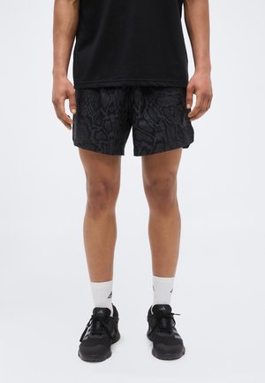 Adulte portant un short noir à motifs, des baskets noires, des chaussettes blanches Adidas et un t-shirt noir, debout devant un fond clair.