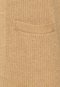 Beige stickad tyg med ett strukturerat mönster, med en horisontell ficka av matchande tyg nära sömmen.