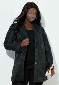 Sort jakke med blomsterprint, skræddersyet pasform, lange ærmer, hakkrave og markante knapdetaljer, båret over en sort top.