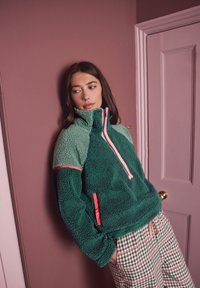 Tumesinine ja heleroheline fliispullover, roosa luku ja detailidega. Omadusteks kõrge krae ja küljetaskud. Asetatud roosa seina vastu.