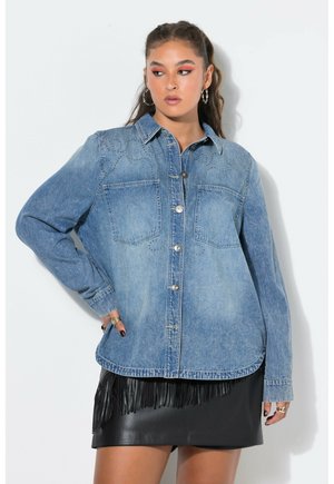 Kvinde iført en blå denimskjorte med knapper og brystlommer samt en sort lædernederdel med frynser, poserer mod en lys ensfarvet baggrund.