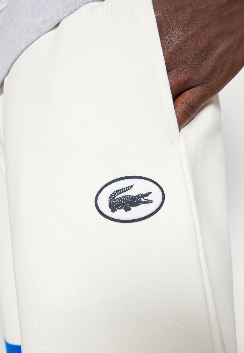 Pantaloni sportivi bianchi con una striscia blu, con un patch ovale blu e bianco del logo Lacoste sulla tasca sinistra.