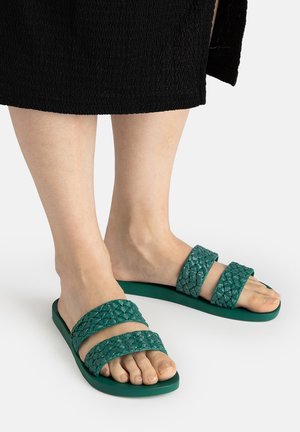 Füße tragen grüne flache Sandalen mit zwei geflochtenen Riemen, stehen auf weißer Oberfläche, untere Beine sichtbar mit schwarzem strukturiertem Rock.