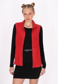 Vesta roșie din fleece cu fermoar pe față și guler înalt, purtată peste o rochie neagră cu mânecă lungă. Prezintă un design ajustat și o textură netedă.