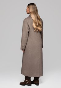 Kvinde med langt blondt hår if�ørt en lang gråbrun frakke og mørkebrune ankelstøvler, stående og vendt sidelæns mod en ensfarvet baggrund.