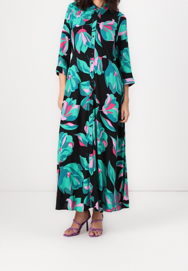 Femme portant une longue robe noire boutonnée avec de grands motifs floraux turquoise et roses et des sandales à talons violet à brides.