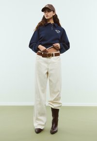 Frau in dunkelblauem, gekürztem Sweatshirt, beigen, hoch-taillierten weit geschnittenen Hosen mit braunem Gürtel, braunen Cowboy-Stiefeln und braunem Hut, der den Saum anpasst.