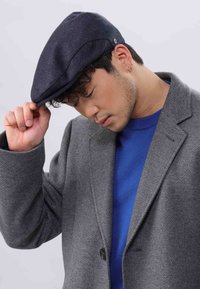 Flat cap in dark blue fabric with smooth texture. Curved bill and subtle logo detail. Worn with a grey coat over a blue sweater.