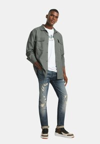 Camicia grigia a maniche lunghe sopra una maglietta bianca, abbinata a jeans blu scoloriti e sneakers multicolori. Design semplice con dettagli della tasca.