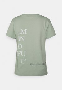 Camiseta de algodón verde claro de manga corta, con un prominente texto blanco que dice ",MINDFUL" verticalmente en la parte posterior, junto con detalles adicionales de la marca.