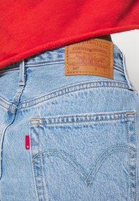 Un jean en denim bleu clair présente une patch en cuir marron avec un logo et des détails en relief, des surpiqûres contrastées et une petite étiquette rouge sur la poche arrière.