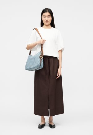 Žena v bielej blúzke a hnedých širokých nohaviciach, drží modrú vzorovanú kabelku cez rameno s koženým remienkom a zlatým príveskom v tvare loga.