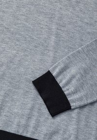 Pull en maille gris avec une manchette noire côtelée. Texture douce avec une finition lisse, montrant des variations subtiles dans la couleur grise.