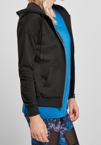 Persona che indossa una felpa nera con zip sopra una maglietta blu e leggings blu-viola con motivo e pannelli in rete, in piedi contro uno sfondo grigio.