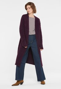 Langer lila fluffiger Cardigan mit offenem Vorderteil, kombiniert mit einem gestreiften Hemd und weiten, dunklen Jeans; das Model trägt braune, Absatz-Ankle-Boots.