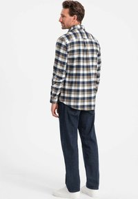 Camicia flanella da uomo a quadri con motivi in blu scuro, marrone e bianco. Maniche lunghe, polsini con bottone e colletto classico. Abbinata a jeans scuri.