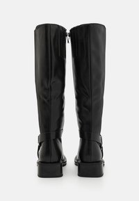 Botas de cuero negro hasta la rodilla con una textura suave, que cuentan con un cierre lateral y una correa alrededor del tobillo. Tacón ancho.