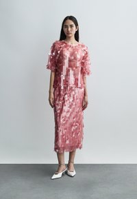 Γυναίκα που φοράει ροζ φόρεμα και τοπ καλυμμένα με μεγάλα παγιέτες, φορώντας λευκές μυτερές γόβες, στέκεται μπροστά σε έναν απλό φωτεινό γκρι τοίχο.