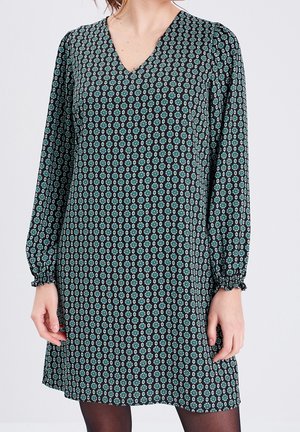 Femme portant une robe noire à manches longues avec des motifs floraux géométriques bleus et blancs et un col en V.