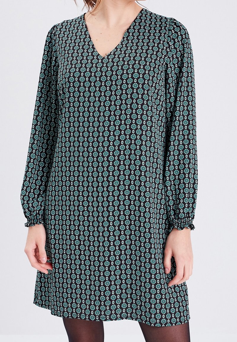 Femme portant une robe noire à manches longues avec des motifs floraux géométriques bleus et blancs et un col en V.