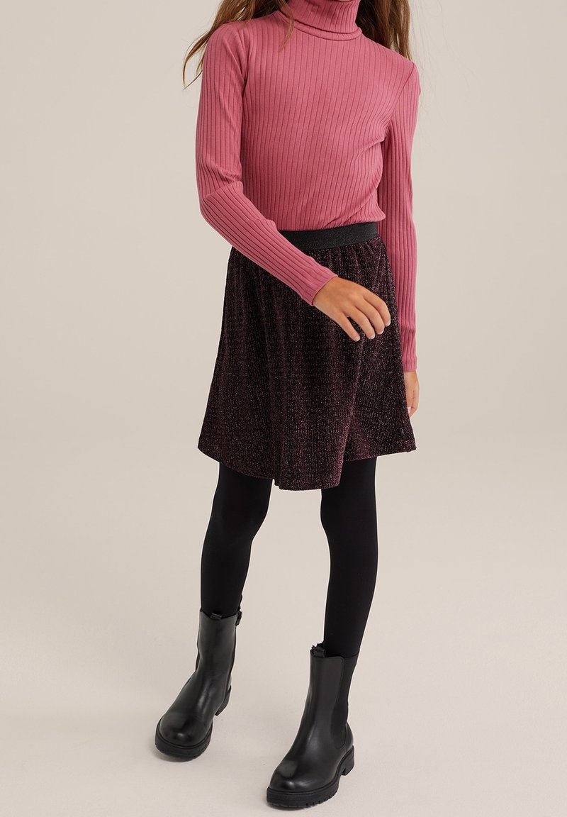 Femme portant un pull à col roulé côtelé rose, une jupe texturée foncée, des leggings noirs et des bottines noires, debout devant un fond uni.