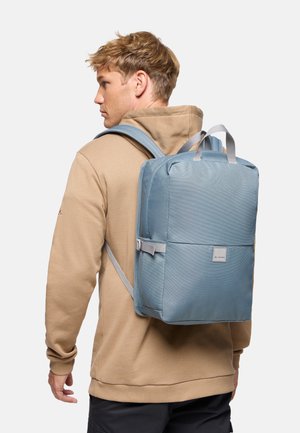 Young man in beige hoodie wearing a light blue rectangular backpack with front logo, adjustable straps, and top carry handles.