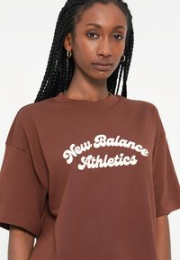 T-shirt oversized castanho em algodão, com um logotipo branco "New Balance Athletics" em uma fonte curva e retro no peito.