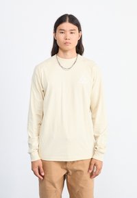 Lys beige langermet skjorte med en trekantet grafikk i hvitt på brystet, sammen med brune bukser. Glatt stoff, avslappet passform.