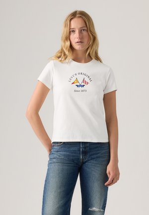 Jeune femme portant un t-shirt blanc Levi's avec des drapeaux nautiques et un jean bleu, debout devant un fond clair uni.