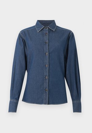 Femme portant un pantalon large foncé et une chemise en jean foncée à boutons, tenant un sac noir avec une bandoulière chaîne, debout devant un fond uni.