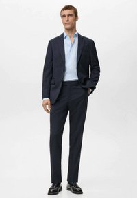 Granatowy garnitur na miarę z jasnoniebieską koszulą. Garnitur ma klapę z nacięciem, dwie przednie kieszenie oraz wąskie spodnie. Dopełnieniem są czarne mokasyny.