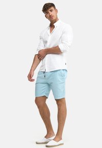 Homme portant une chemise blanche à manches longues avec boutons, un short bleu clair et des chaussures blanches sans lacets, ajustant sa manche gauche sur un fond uni.