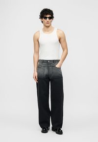 Hombre con gafas de sol negras, camiseta sin mangas blanca y vaqueros de pierna ancha bicolor en negro y gris, de pie con una mano en el bolsillo.