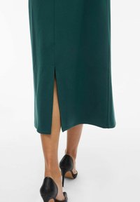 Groene midi-jurk met een zijsplit, gemaakt van zachte stof. Een paar zwarte hoge hakken zijn zichtbaar aan de onderkant.