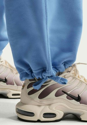 Sneakers Nike beige chiaro e marroni indossate con pantaloni della tuta blu legati alla caviglia, mostrando i dettagli della scarpa e il design della suola.