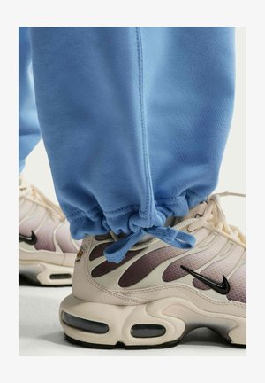 Baskets Nike beige clair et marron portées avec un pantalon de survêtement bleu noué à la cheville, montrant les détails de la chaussure et le design de la semelle.