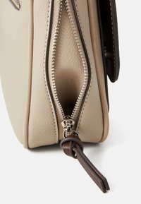 Borsa in pelle beige con finitura texturizzata, caratterizzata da una cerniera argentata e un'acca in pelle marrone vicino al cursore. Vista laterale dell'apertura.