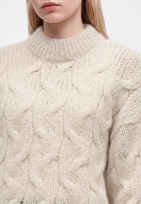 Gros plan d'une personne portant un pull en maille torsadée douce de couleur crème avec un col montant côtelé.