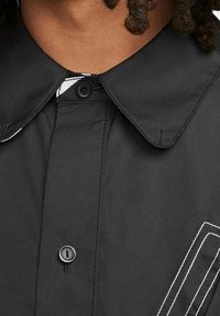 Chemise noire avec un grand col, une fermeture à boutons et des surpiqûres blanches en accent sur le côté gauche. Le tissu semble lisse et résistant.
