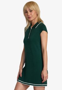 Strick-Polo-Kleid in Dunkelgrün mit kurzen Ärmeln, weißem Kragen und Saum-Akzenten sowie einer figurbetonten Form. Die strukturierte Stoffqualität sorgt für visuelles Interesse.