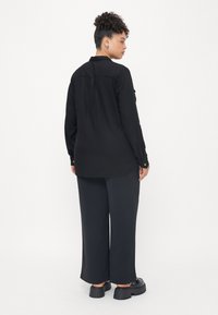 Chemise noire à manches longues avec un col, dotée d'un pli au dos, associée à un pantalon noir ample et des chaussures à plateforme noires.