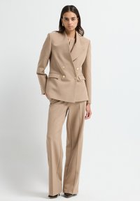 Frau, die auf einem weißen Hintergrund steht und einen beigefarbenen, doppelt geknöpften Blazer mit goldenen Knöpfen, passende weit geschnittene Hose, schwarze Schuhe und goldene Ohrringe trägt.