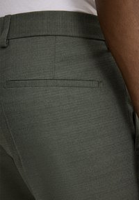 Pantaloni sagomati verde scuro con una texture liscia, caratterizzati da un taglio dritto e una tasca posteriore singola senza elementi hardware visibili.