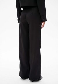 Hoge zwarte broek met wijde pijpen, gemaakt van een soepele stof. Kenmerkt zich door minimale naden en onopvallende zijzakken. Schoeisel gedeeltelijk zichtbaar.