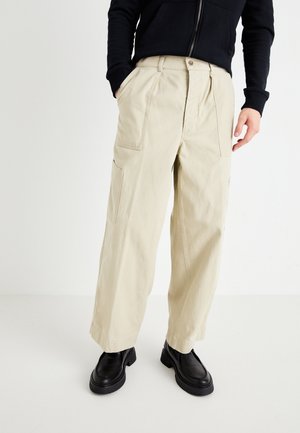 Beige cotton cargo pants with wide legs, front pleats, and side pockets. Paired with black chunky shoes. Simple, functional design.