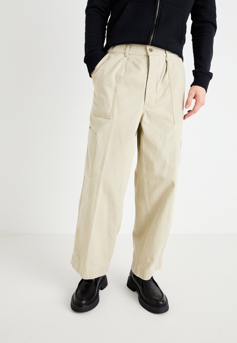 Beige cotton cargo pants with wide legs, front pleats, and side pockets. Paired with black chunky shoes. Simple, functional design.