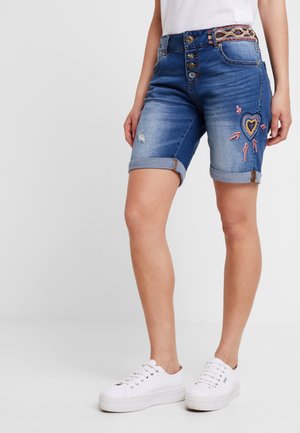 Femme portant un short en denim bleu avec une broderie en forme de cœur et une ceinture à motifs, assorti à des baskets blanches.