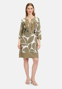 Khakifärgad och krämfärgad knälång klänning med bladmönster, v-ringning och trekvartsärmar, har metalliska detaljer på framsidan.