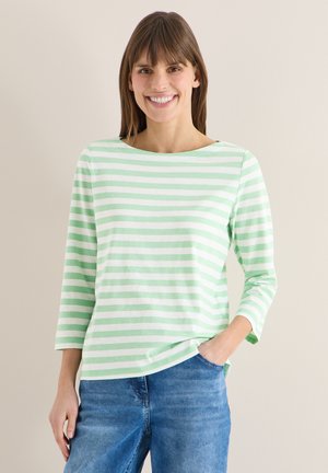 Lächelnde Frau, die ein langärmeliges Shirt mit hellgrünen und weißen Streifen und blaue Jeans trägt, mit einer Hand in der Tasche, stehend vor einer schlichten Wand.